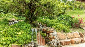 Un jardín con una pequeña cascada, rodeado de abundante follaje y un muro de piedra.