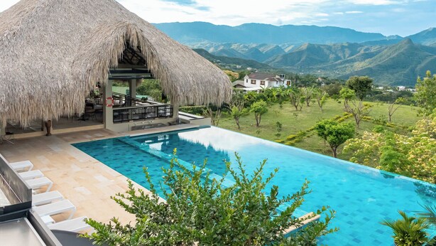 Zona junto a la piscina con una estructura de techo de paja, una piscina azul cristalina y vistas a montañas distantes.