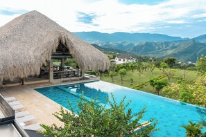 Uma área à beira da piscina com estrutura de telhado de palha, piscina azul límpida e vista para montanhas distantes.