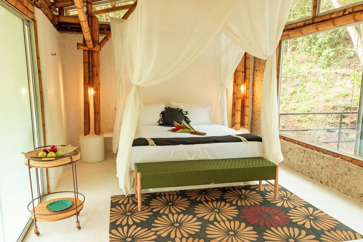 A room with a bed, a small table with fruit, and a view of greenery outside.