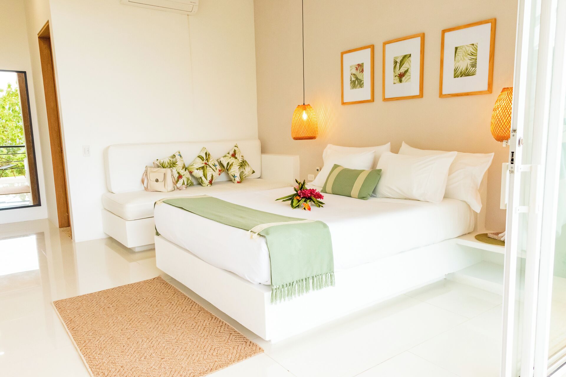 A modern bedroom with a large bed, a sofa, and a small table. There are framed pictures on the wall and a window with a view of greenery.