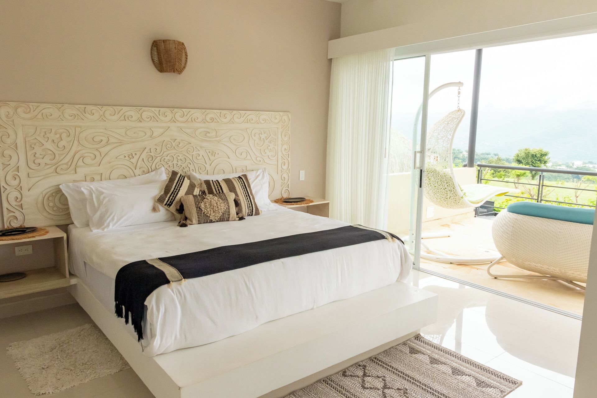 A spacious bedroom with a large bed, a decorative headboard, and a view of a balcony with outdoor seating.
