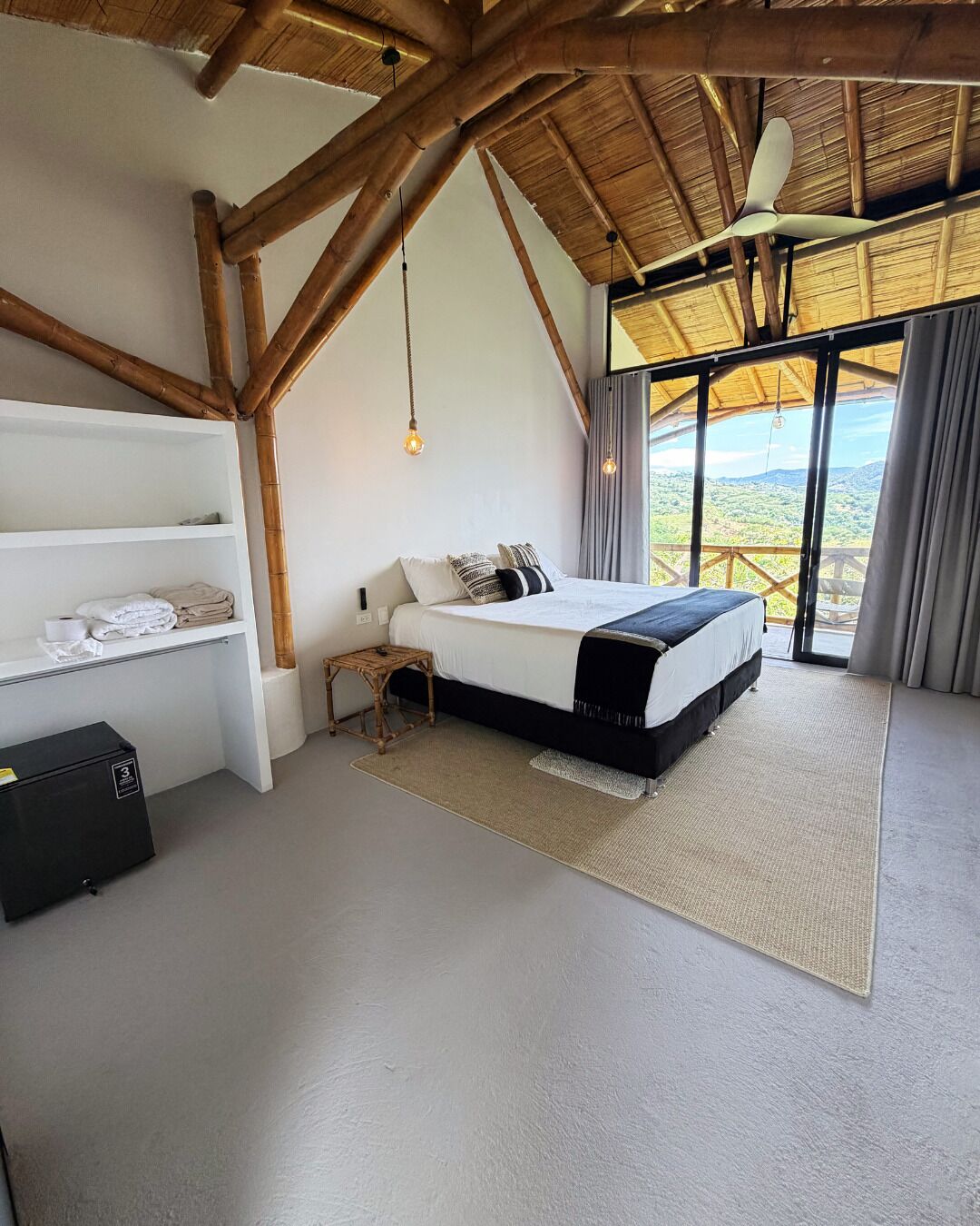 Une chambre à coucher avec un lit, une table de chevet, une étagère avec des serviettes et une vue sur l’extérieur.