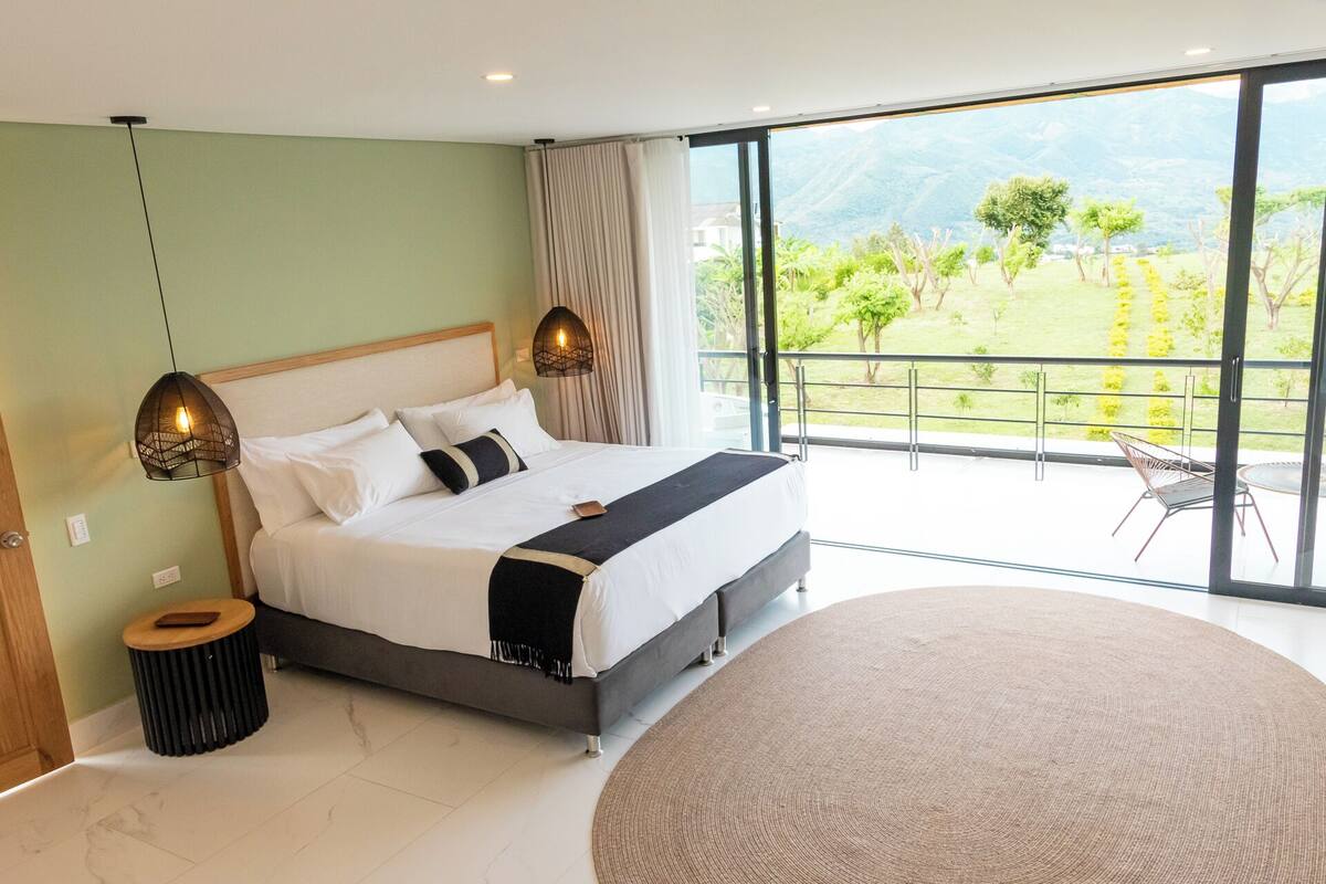 A modern bedroom with a large bed, two pendant lights, a round rug, and a balcony with a view of greenery and mountains.