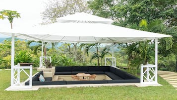 Un espacio de estar al aire libre con techo, toldo blanco, cojines negros y mobiliario de madera.