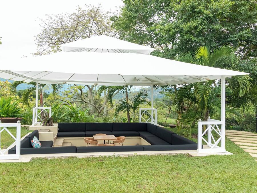 Un espacio de estar al aire libre con techo, toldo blanco, cojines negros y mobiliario de madera.