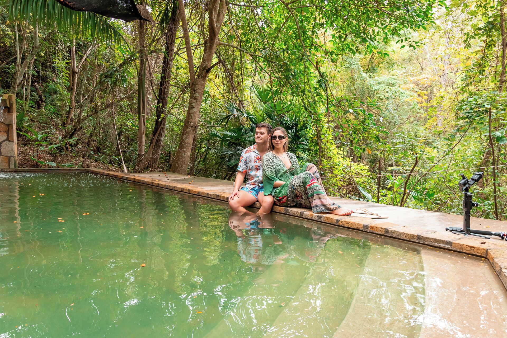 A couple sitting by a small pool surrounded by dense greenery.