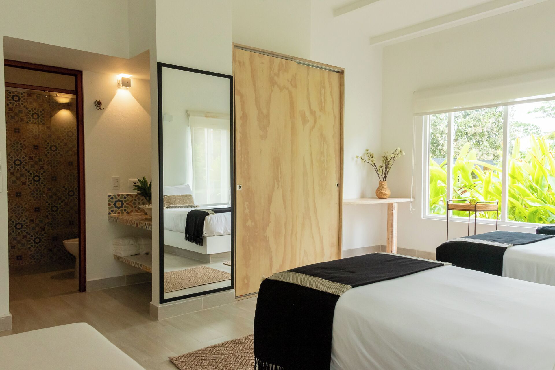 A bedroom with a large mirror, a wooden wardrobe, a bedside table with a vase, and a window with a view of greenery.