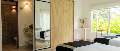 A bedroom with a large mirror, a wooden wardrobe, a bedside table with a vase, and a window with a view of greenery.