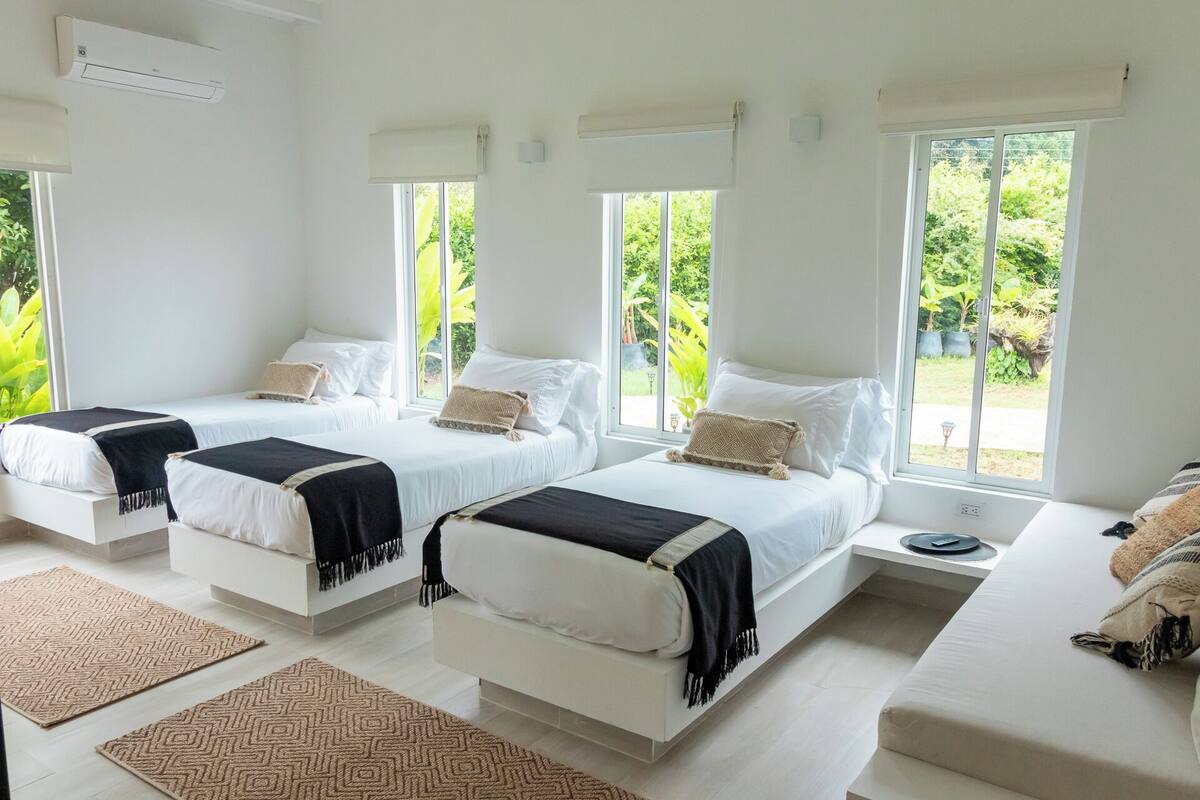 A hotel room with three beds, large windows, and a view of greenery.