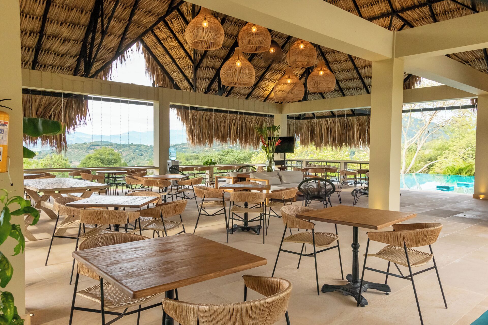 A restaurant with thatched ceilings, wicker furniture, and a view of greenery and mountains.