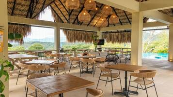 A restaurant with thatched ceilings, wicker furniture, and a view of greenery and mountains.