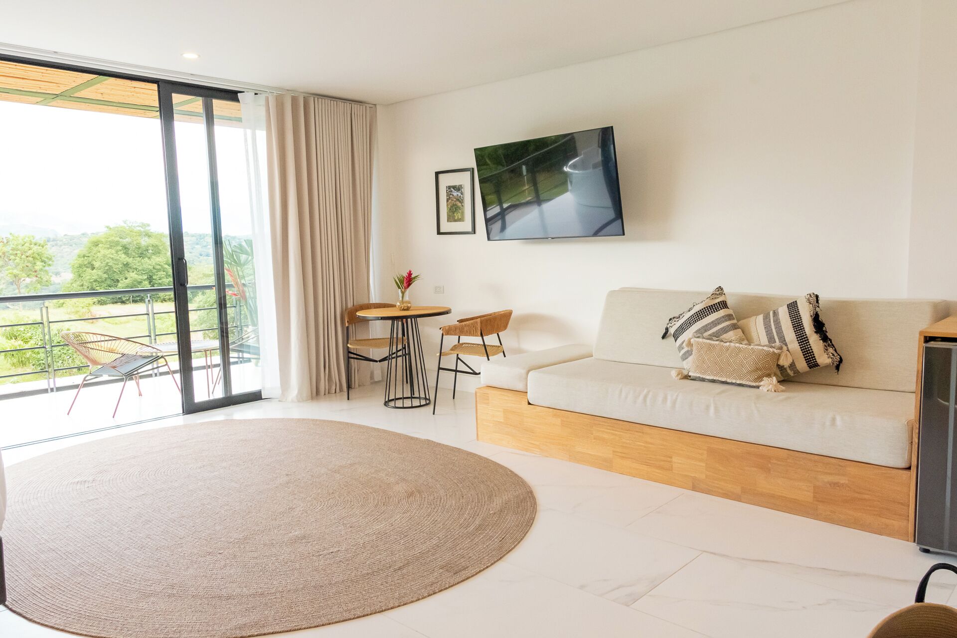 A modern living room with a sofa, a round rug, a dining table with chairs, and a TV mounted on the wall.