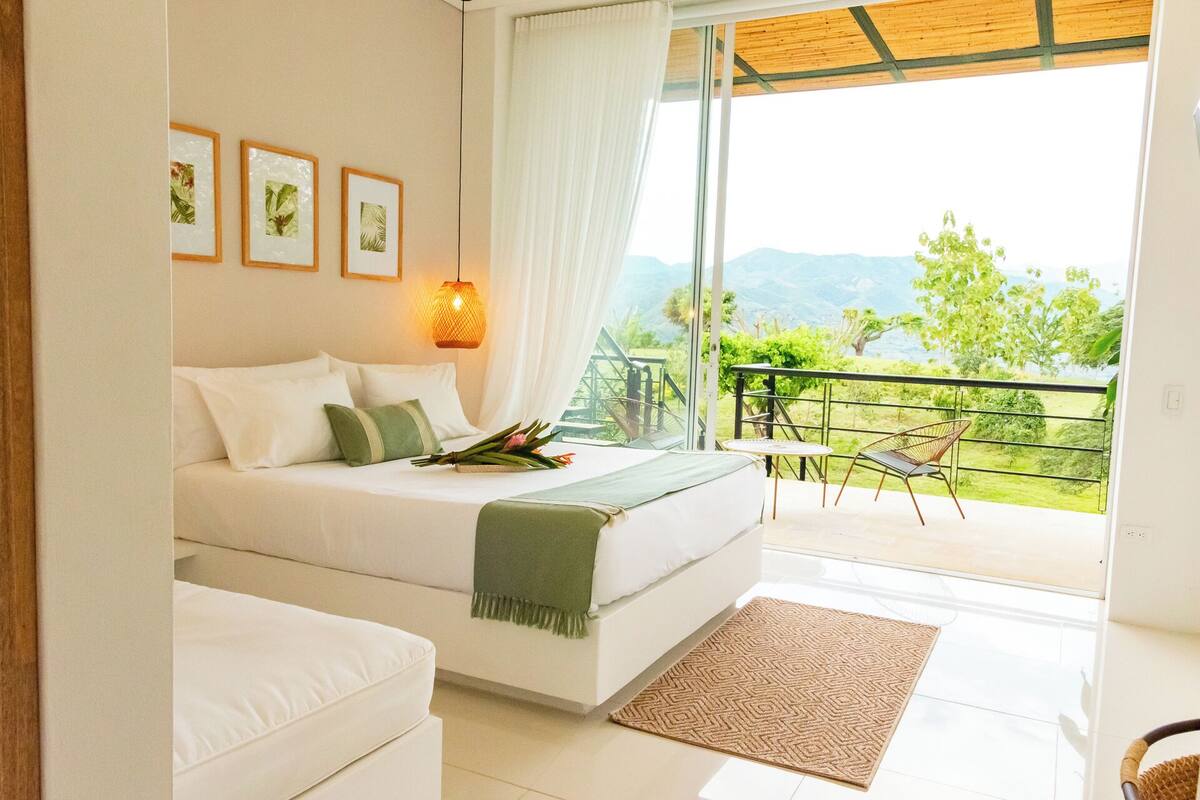 A bedroom with a bed, a bedside table, a lamp, and a view of a balcony with a railing and greenery.