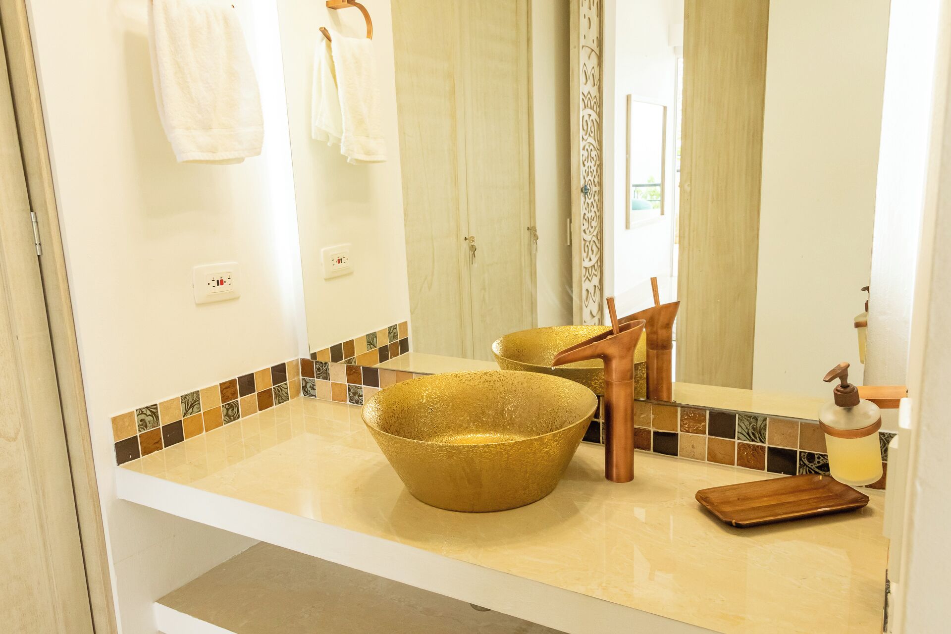 A bathroom with a large gold bowl sink, a wooden chair, and a mirror reflecting the room.