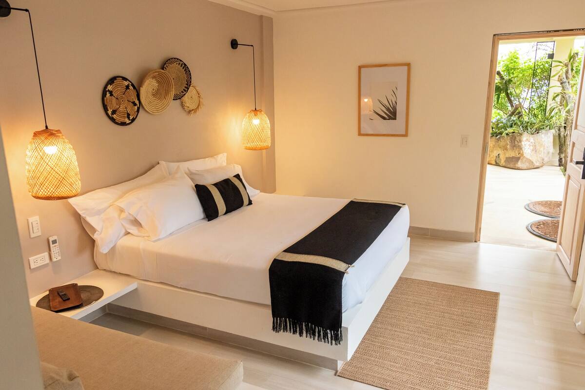 A neatly arranged bedroom with a bed, bedside lamps, a wall-mounted phone, and a door leading to an outdoor area with plants.