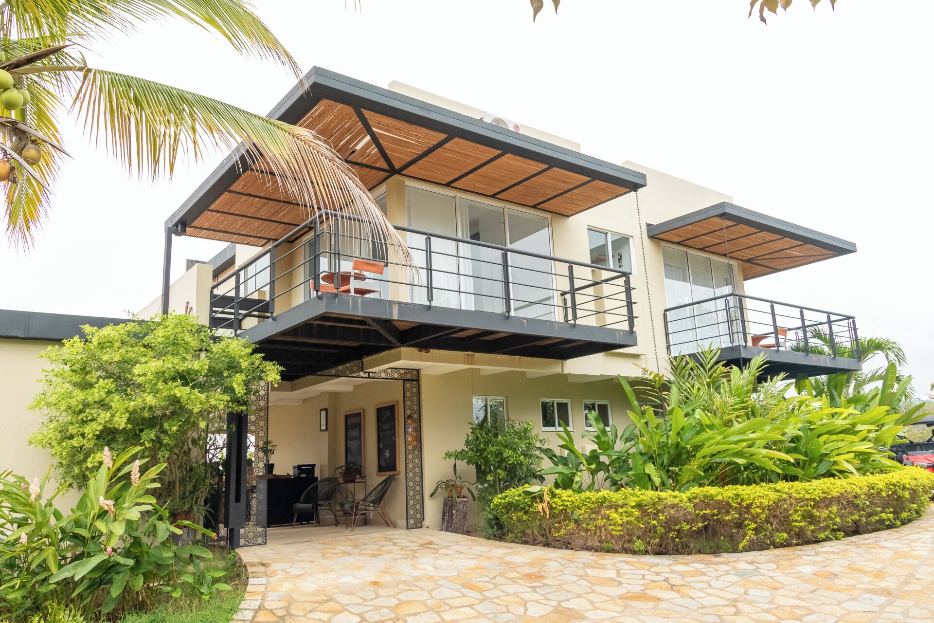 A modern two-story house with a balcony, a stone-paved entrance, and lush greenery.