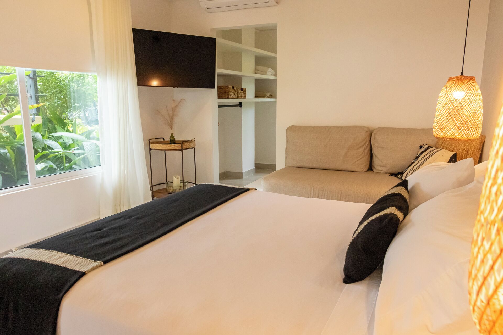 A bedroom with a bed, a bedside table, a hanging light fixture, and a window with a view of greenery.