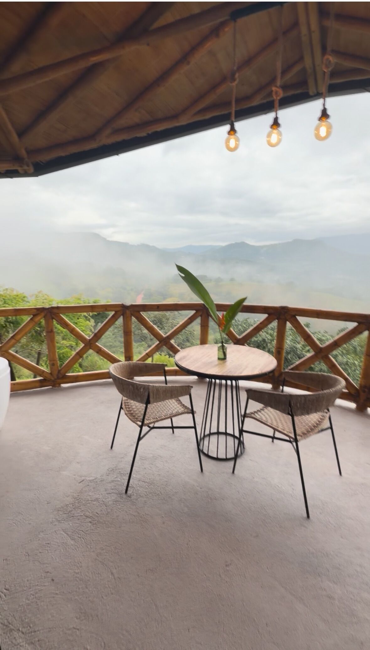 Una mesa redonda con cuatro sillas de mimbre, una pequeña planta en maceta y lámparas colgantes en un balcón con barandilla de madera, con vistas a un paisaje montañoso.