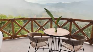 Una mesa redonda con cuatro sillas de mimbre, una pequeña planta en maceta y lámparas colgantes en un balcón con barandilla de madera, con vistas a un paisaje montañoso.