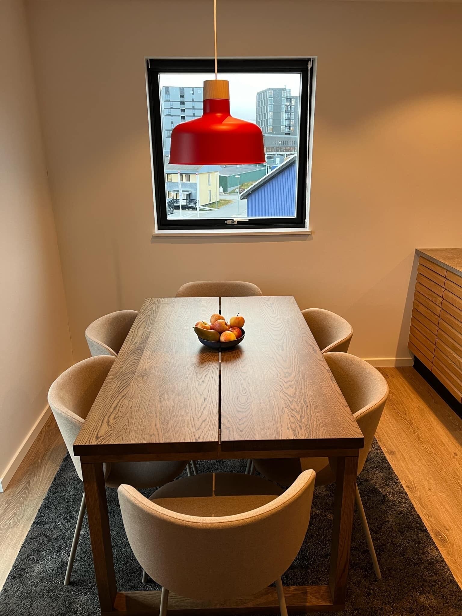 A dining area with a wooden table, four chairs, a bowl of fruit, and a red pendant light above a window with a city view.