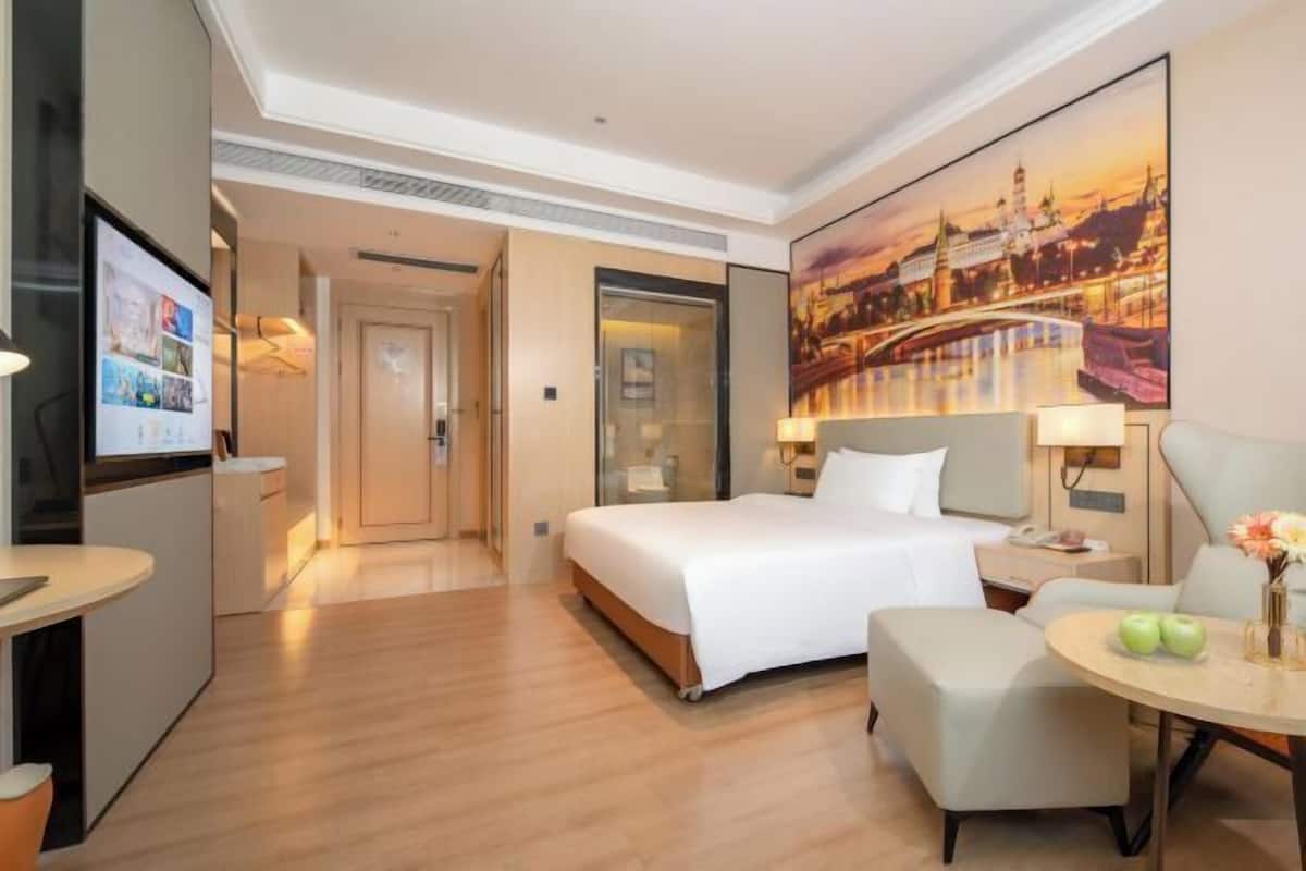 A hotel room with a bed, a desk, a chair, a table with apples, a TV, and a painting of a cityscape.