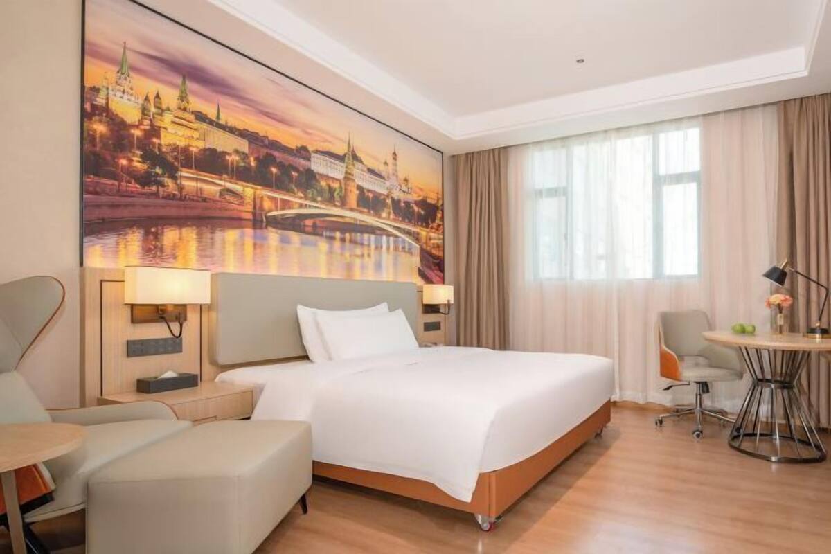 A hotel room with a large bed, a desk, a chair, a small table, and a painting of a cityscape.