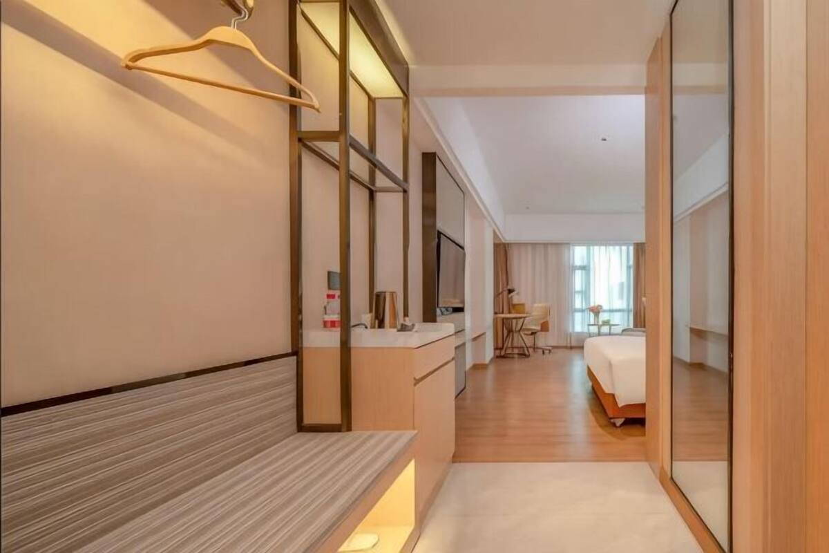 A hotel room with a bed, bedside table, and a mirror. There is a bench along the wall and a desk with a chair.