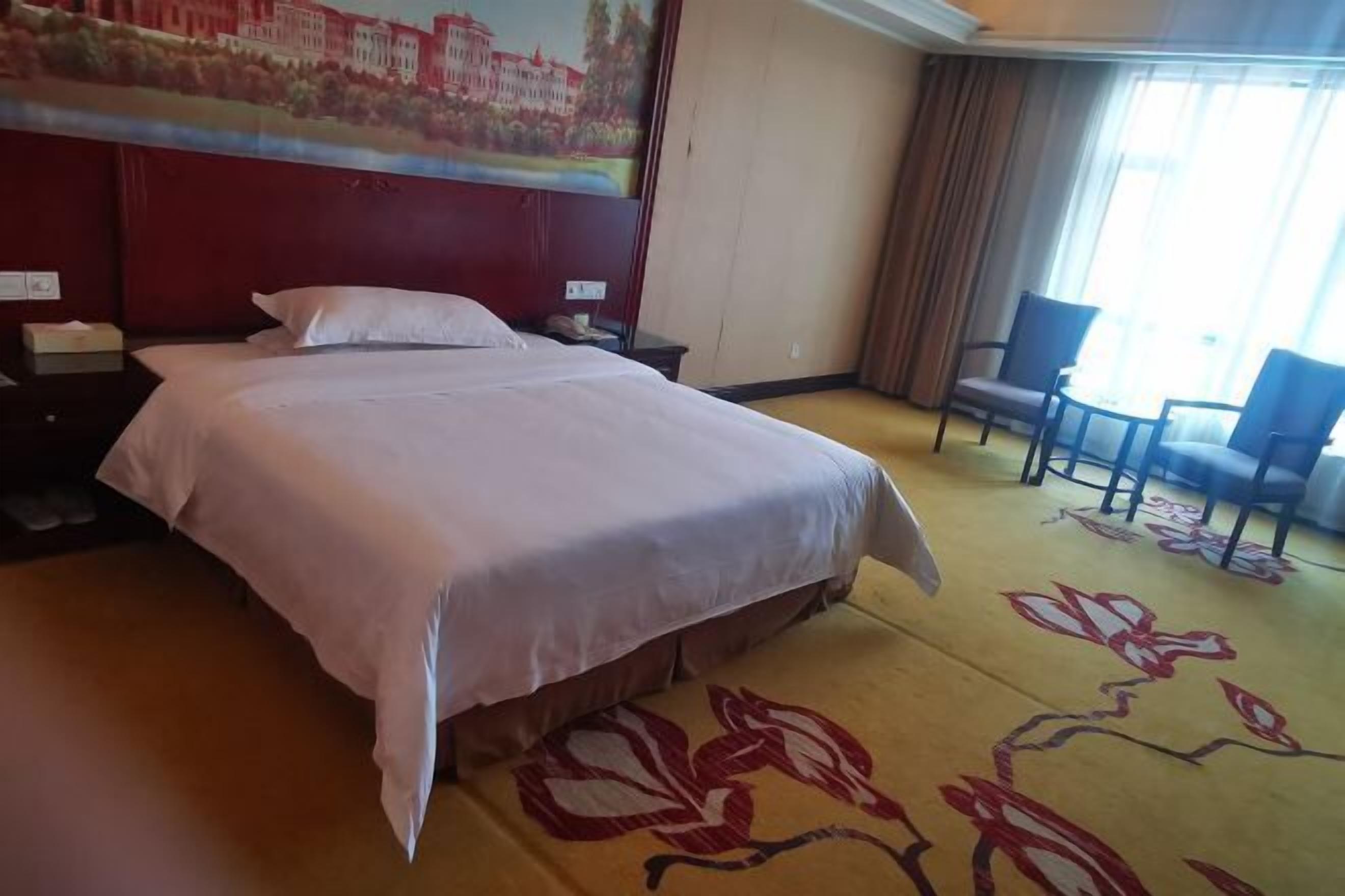 A hotel room with a large bed, a headboard with a painting, a carpet with a floral pattern, and two chairs.