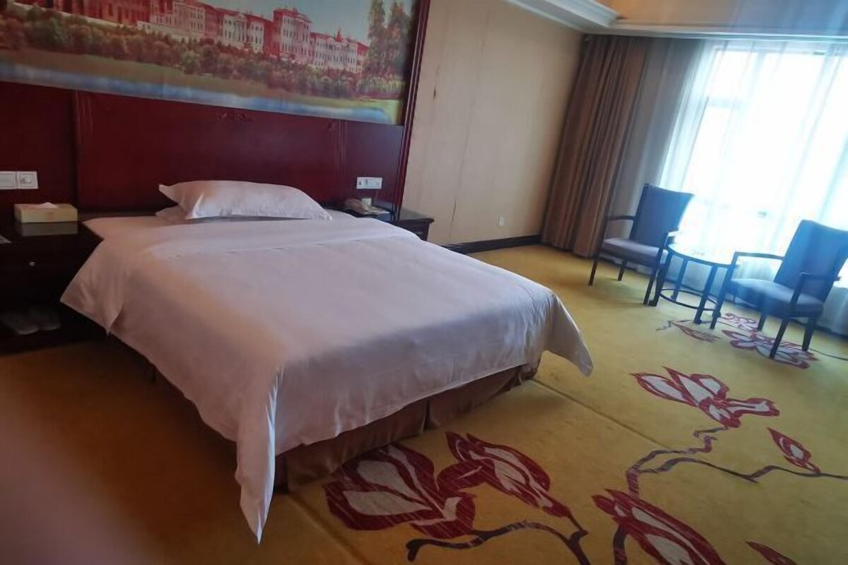 A hotel room with a large bed, a headboard with a painting, a carpet with a floral pattern, and two chairs.
