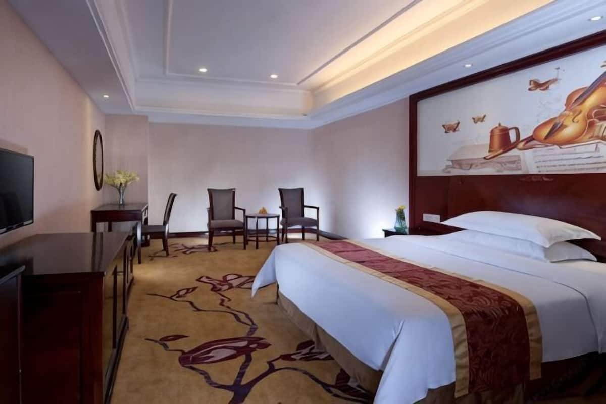 A hotel room with a large bed, a television, two armchairs, a small table, and a decorative wall piece.