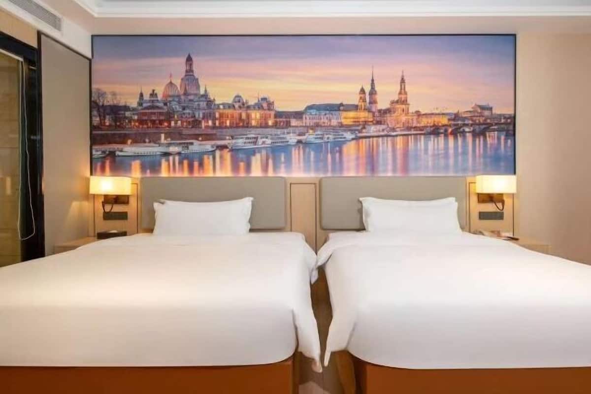 A hotel room with two beds, a large cityscape painting on the wall, and two bedside lamps.