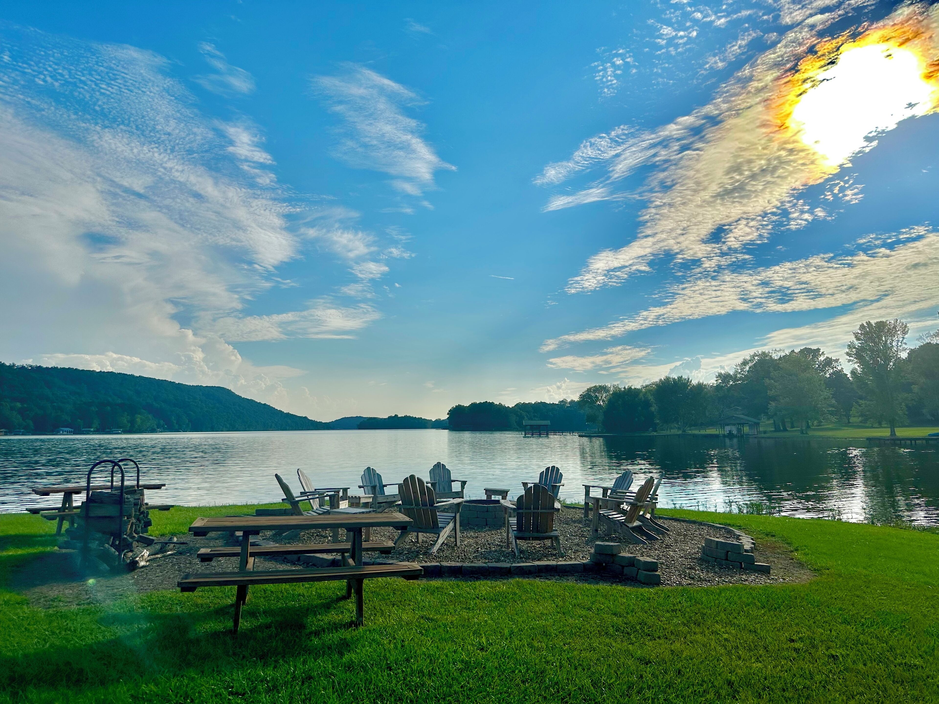 A lakeside picnic area with Adirondack chairs and a wooden table, surrounded by a grassy area and a dock extending into the water.