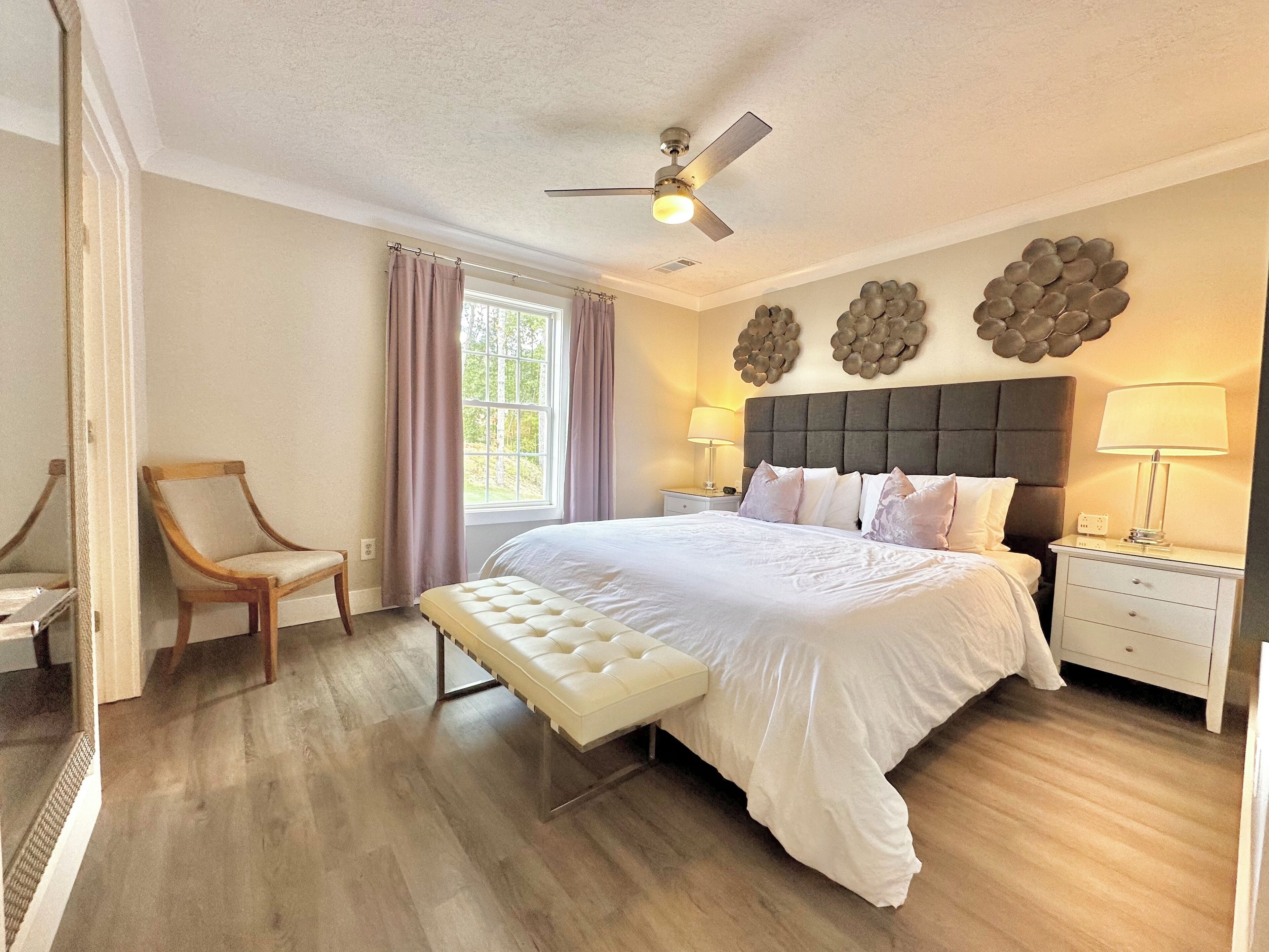 Une chambre à coucher avec un grand lit, un banc, une chaise, un ventilateur de plafond et une fenêtre avec des rideaux.