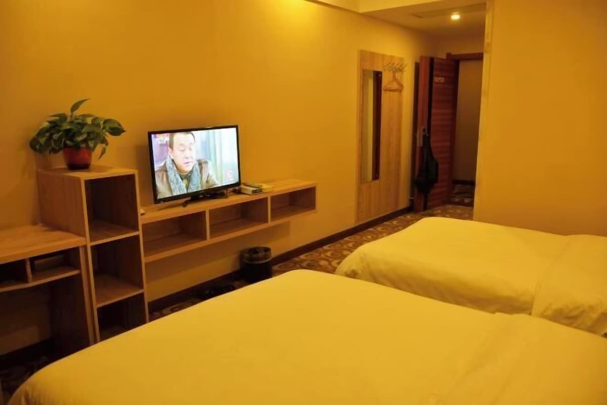 A hotel room with two beds, a TV on a wooden shelf, a potted plant, and a wooden door.