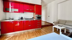 Una cucina moderna con mobili rossi, piano cottura a gas e forno. C'è un piano di lavoro bianco, un lavello e un piccolo tavolo bianco. Nell'area soggiorno, un divano color beige e una sedia bianca.