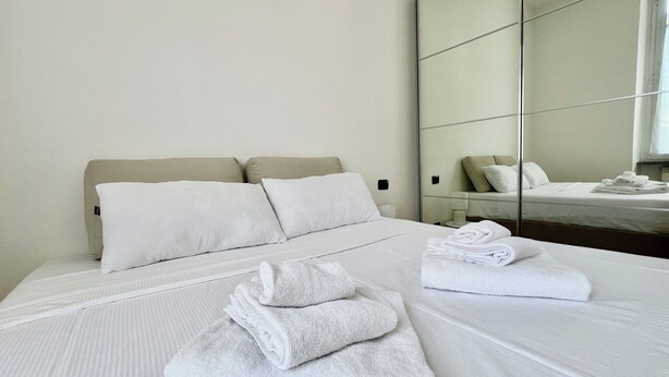 Camera d'albergo con un letto grande, due cuscini, un asciugamano piegato con cura e uno specchio che riflette il letto e una finestra.