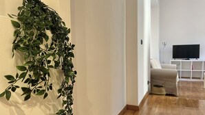 Un soggiorno con pavimento in legno, un divano bianco, una televisione su un mobile bianco e un’ampia pianta verde sulla parete.