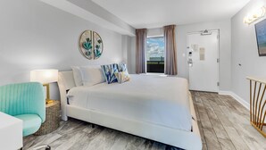 A modern hotel room with a large bed, a bedside table with a lamp, a view of the city through a window, and a door with a sign. - Pristine Oceanfront Suite with Enticing Views (Myrtle Beach)