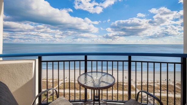 A balcony with a glass table and chairs, overlooking a beach with a clear sky and clouds. - Pristine Oceanfront Suite with Enticing Views (Myrtle Beach)