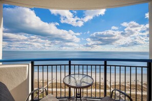 A balcony with a glass table and chairs, overlooking a beach with a clear sky and clouds. - Pristine Oceanfront Suite with Enticing Views (Myrtle Beach)
