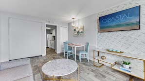 A modern interior with a dining area, a TV displaying 'COASTAL PROPERTIES', and a herringbone-patterned wall. - Pristine Oceanfront Suite with Enticing Views (Myrtle Beach)