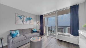 A modern living room with a grey sofa, a round coffee table, and a large sliding glass door leading to a balcony with a view of the sea.