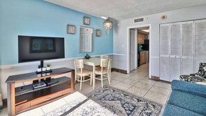 A living room with a TV, dining table, chairs, and a sofa. There is a kitchen in the background with cabinets and a refrigerator. - Unforgettable Beachfront Bliss (Myrtle Beach)