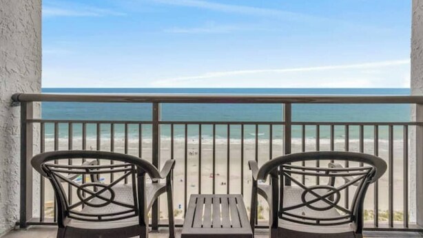 Ein Balkon mit zwei Stühlen und einem kleinen Tisch, der Blick auf einen Strand und das klare Meer gewährt.