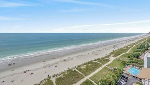 Ein Strand mit freiem Blick auf den Ozean und einem Poolbereich mit Liegestühlen und Sonnenschirmen.