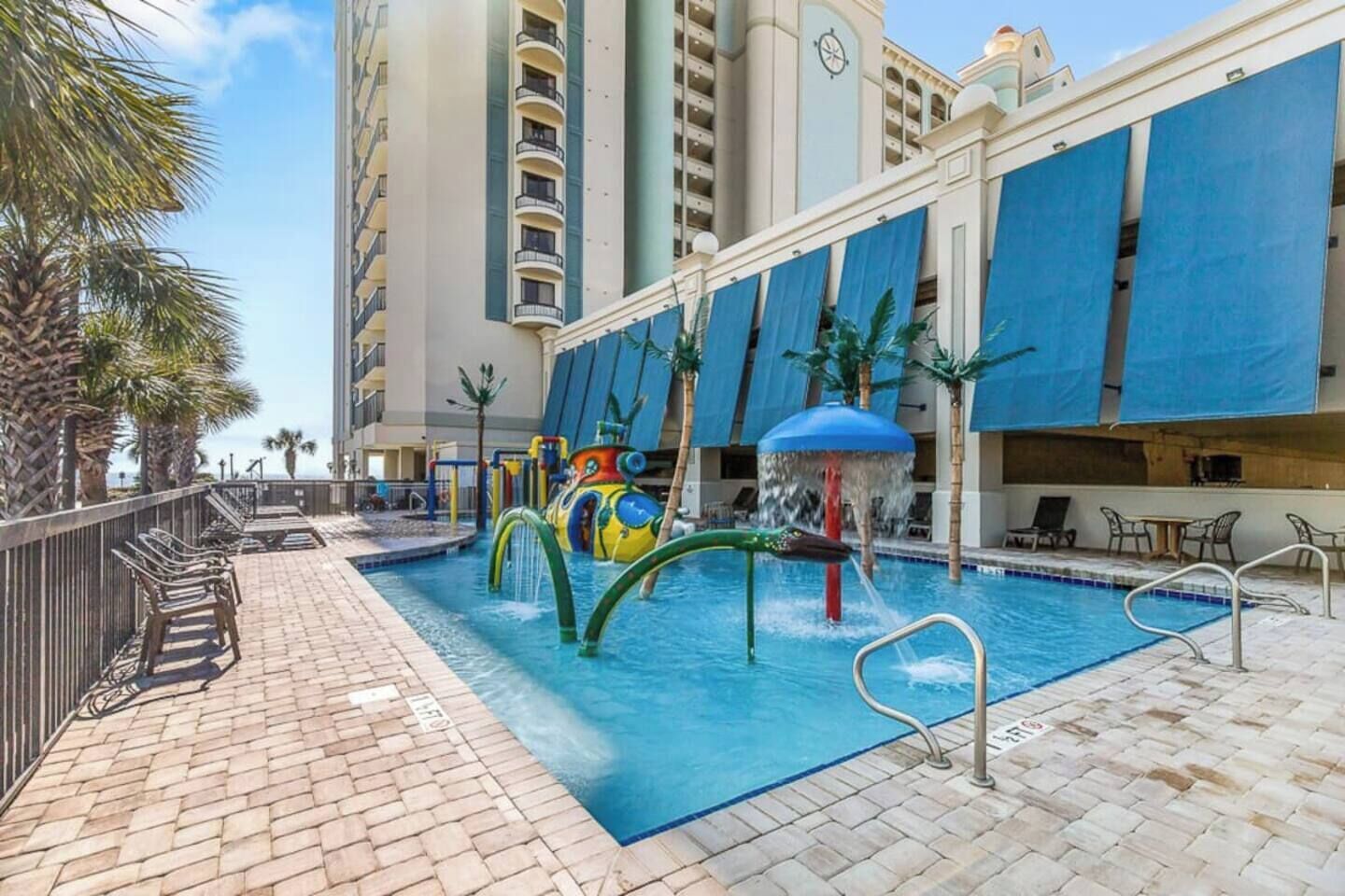 A swimming pool with a water play area, surrounded by a brick deck and palm trees.
