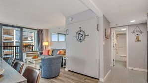 A modern living room with a sofa, a coffee table, and a dining area with chairs. There is a large window with a view of balconies. - Escape to the Ocean~ Oceanview Suite @CompassCove (Myrtle Beach)
