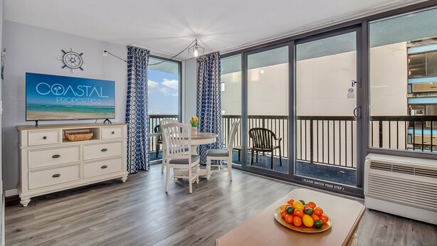 A spacious balcony with a view of the sea, a dining area with a table and chairs, and a television displaying 'COASTAL PROPERTIES'. - Escape to the Ocean~ Oceanview Suite @CompassCove (Myrtle Beach)