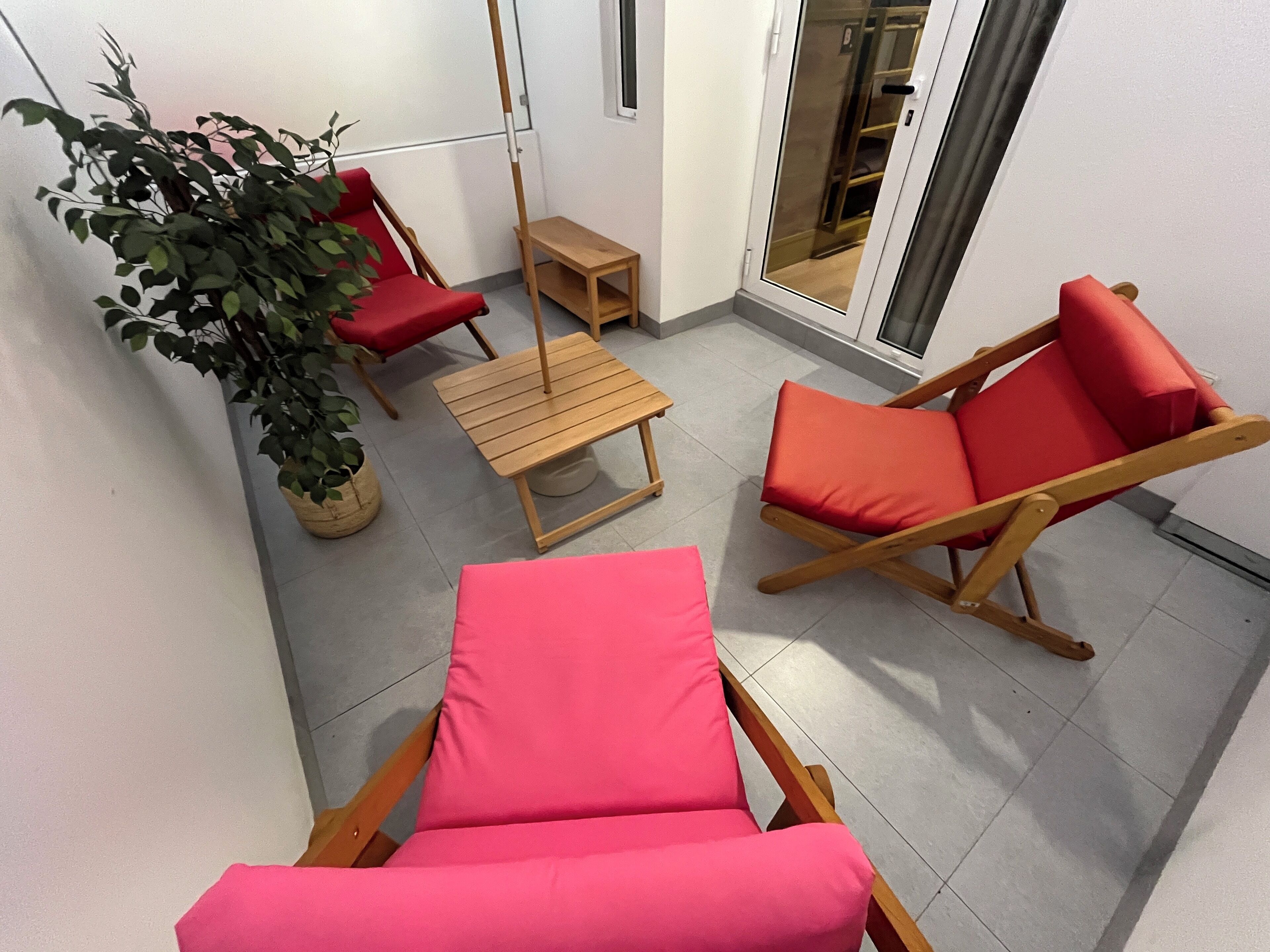 A small balcony with two red lounge chairs, a wooden table, and a potted plant.